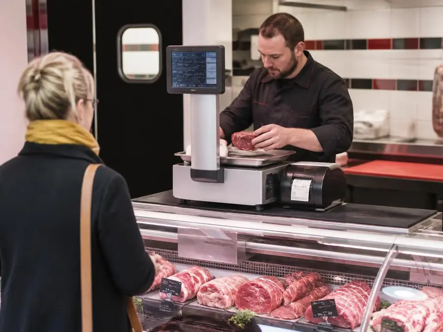 Boucher qui pèse une pièce de viande sur une balance connectée avec imprimante d'étiquettes devant sa cliente